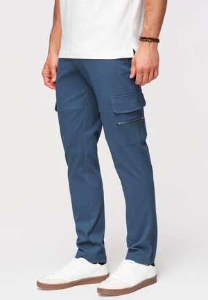 Mężczyzna w białych trampkach, niebieskich spodniach cargo z kieszeniami na suwak i patkę oraz białej koszuli, stojący na jednolitym tle.