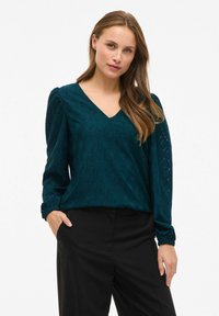 Teal blouse met lange mouwen en een V-hals, met een gestructureerd ruitpatroon, gathered mouwen en een losse pasvorm, gecombineerd met zwarte broek.