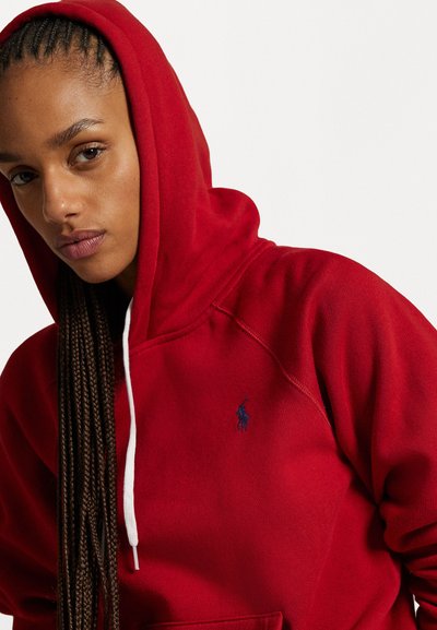 Rød hettegenser laget av myk stoff med hvit snor. Har et logoemblem på venstre bryst og flettet hår synlig.