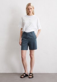 White cotton t-shirt featuring a small logo, paired with dark blue shorts and black sandals with a gold accent buckle. Minimal design.