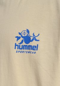 Beiges Baumwoll-T-Shirt mit einem blauen Hummel Sportswear-Logo, das einen Comic-Charakter und ein Pflanzendesign zeigt, deutlich auf der Vorderseite platziert.