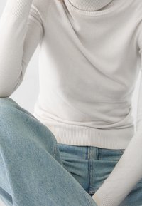 Suéter de cuello vuelto de crema hecho de un tejido suave, con puños y dobladillo acanalados, combinado con jeans de mezclilla azul claro. Detalle texturizado visible.