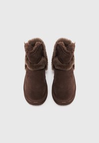 Cizme ankle din suede maro, cu o căptușeală moale și pufoasă, vârf rotund și un design cu mansetă pliată. Talpă plată cu detalii de cusătură.
