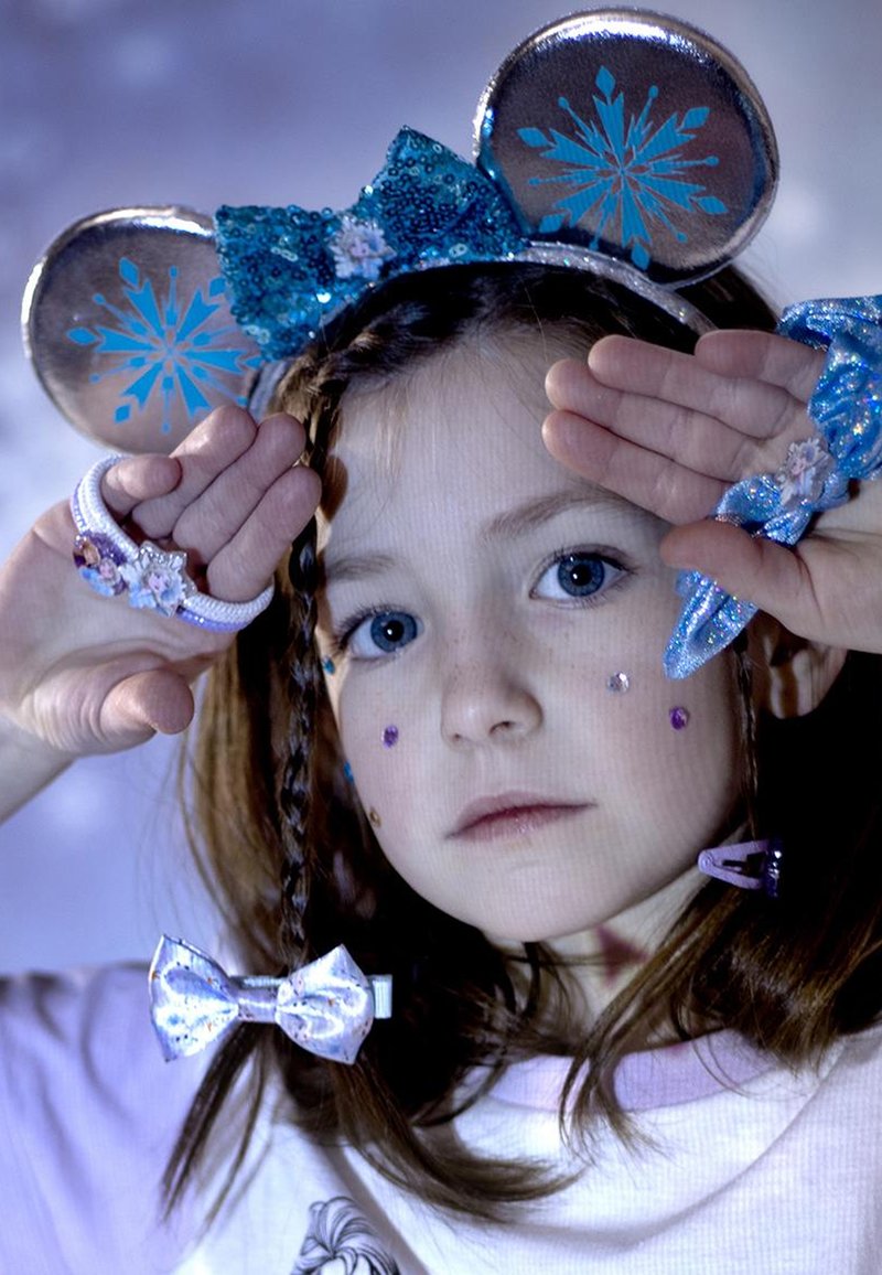 Orecchie di topo blu con paillettes e motivi di fiocchi di neve, capelli intrecciati e accessori scintillanti. Indossa una maglietta rosa con una stampa di personaggi.