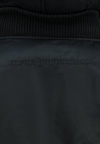 Czarna kurtka z tyłu z żebrowanym kołnierzem i subtelnym czarnym haftowanym tekstem "ALPHA INDUSTRIES" na górnej części pleców.