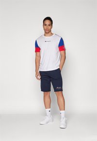 Camiseta blanca con mangas azules y rojas, logo en el pecho, combinada con pantalones cortos color marino con logo, usada con zapatillas y calcetines blancos.
