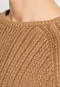 Gros plan sur le cou et l'épaule d'une personne portant un pull tricoté marron clair texturé avec un col rond côtelé.