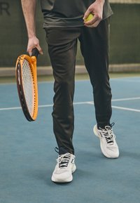 Schwarze Sporthose, weiße Tennisschuhe mit schwarzen Schnürsenkeln und gelben Akzenten sowie ein Tennisschläger mit einem orangefarbenen Griff und schwarzem Saiten.