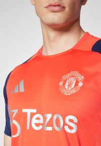 Sarkans īsām piedurknēm sporta krekls ar zilām akcentiem, priekšpusē ir Mančestras United simbolika un "Tezos" sudraba krāsā.
