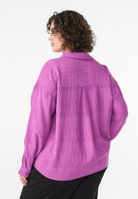 Košeľa s dlhými rukávmi v žiarivej fialovej farbe s textúrovaným vzorom, vybavená golierom a zapínaním na gombíky na manžetách, nosená cez čierne nohavice.