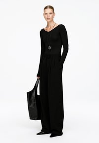 Frau mit zurückgegelten Haaren trägt ein enganliegendes schwarzes Langarmoberteil, weite Hosen, spitze Schuhe, eine Halskette und hält eine große schwarze Tragetasche.