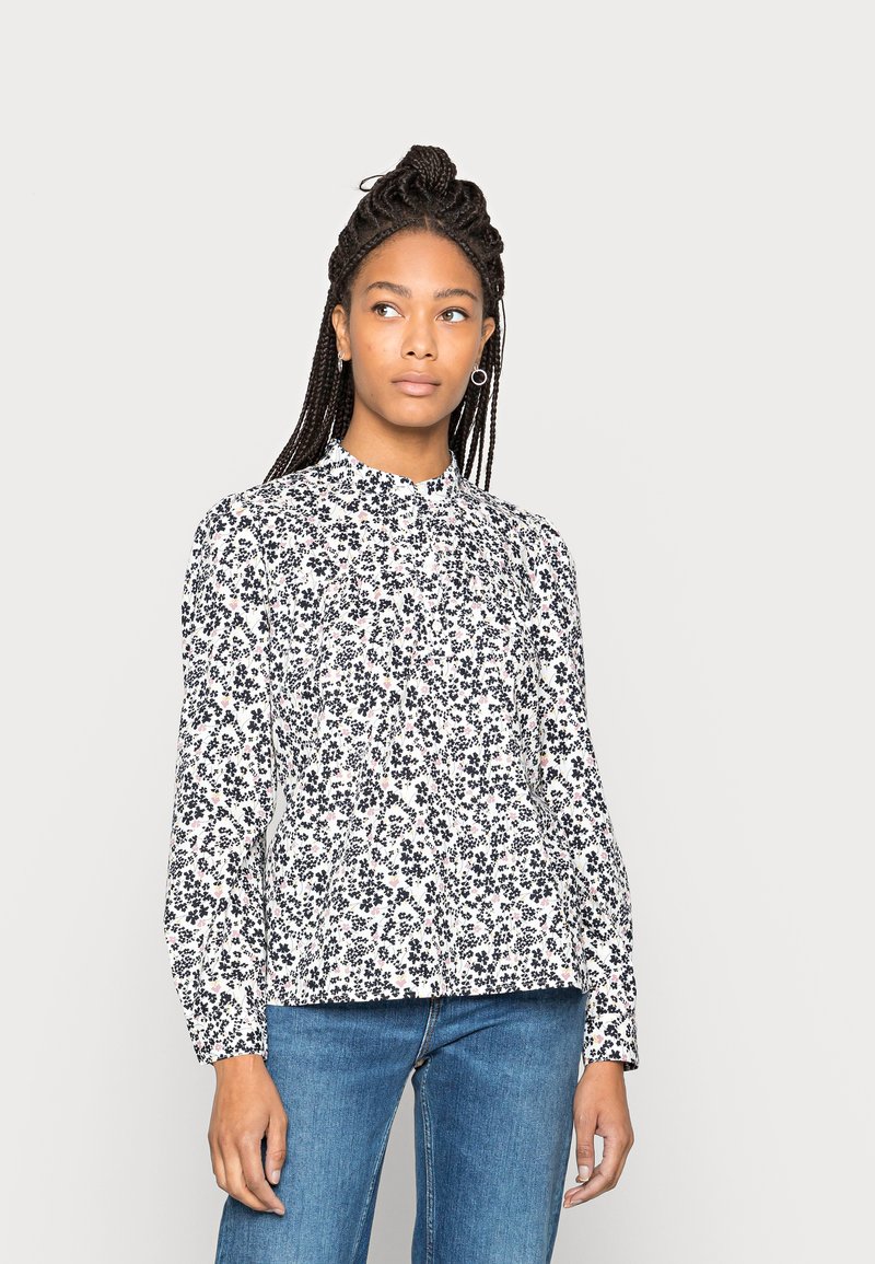 Blusa branca de mangas longas com pequeno padrão floral preto, colarinho alto e detalhes franzidos. Combinada com jeans de ganga azul.