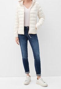 Cremefarvet pufferjakke med horisontalt kviltet design, parret med mørkeblå jeans og hvide sneakers. Enkel V-hals t-shirt under.
