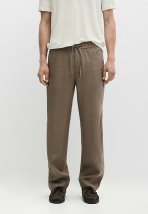 Uomo che indossa pantaloni di lino marrone con coulisse, una camicia crema a maniche corte e scarpe casual marrone scuro, in piedi davanti a uno sfondo semplice.
