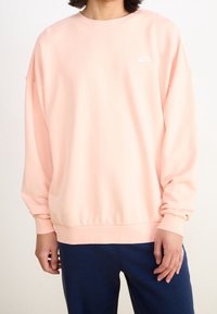 Ljusrosa sweatshirt av mjukt tyg, med rund halsringning, sänkta axlar och en liten vit logga på bröstet.