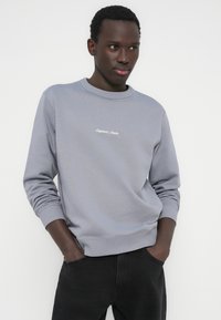 Ανοιχτό γκρι φούτερ με στρογγυλή λαιμόκοψη και ριμπ μανσέτες. Διαθέτει διακριτικό κεντητό κείμενο στο στήθος. Φοράει σκούρο παντελόνι.