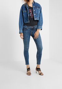 Woman wearing cropped blue denim jacket, black graphic tee, skinny blue jeans with frayed hems, and black strappy high-heeled sandals.