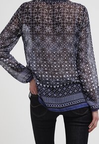 Przezroczysta granatowa bluzka z misternym jasnoniebieskim wzorem, z długimi rękawami, zaokrąglonym dołem i warstwowym wyglądem na ciemnych dżinsach.