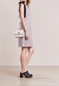 Žena v bezrukávových bílých šatech s vertikálními červenými pruhy, černými lodičkami na vysokém podpatku a bílou kabelkou přes rameno se zlatým řetízkem.