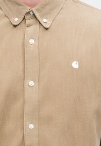 Καφέ μπεζ πουκάμισο από κορντούρο με γιακά που κλείνει με κουμπιά, διαθέτει λευκά κουμπιά και μικρό λευκό λογότυπο στο στήθος. Κατακόρυφη ριμπ υφή.