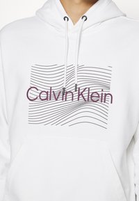 Sweat à capuche blanc avec des lignes ondulées noires et le texte "Calvin Klein" violet sur le devant, doté de cordons de serrage et d'une poche avant.