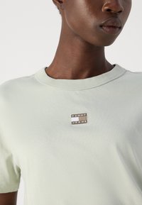 Šviesiai žalias medvilninis marškinėlis su apvaliu iškirpimu ir trumpomis rankovėmis. Jame yra sidabrinė "TOMMY JEANS" logotipo etikete ant krūtinės.