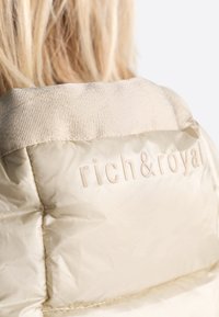 Béžová bunda s péřovou výplní, která má texturovaný límec, prošívané detaily a embosované jméno značky "rich&royal" na zadní straně.