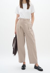 Pískové kalhoty s širokými nohavicemi a skladbami, zkombinované s bílým tričkem a černými mokasíny. V jedné ruce je držena hnědá plátěná taška.