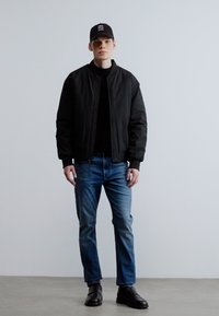 Μαύρο bomber μπουφάν πάνω από μαύρο πουλόβερ, μπλε τζιν και μαύρα παπούτσια. Το μοντέλο φοράει μαύρο καπέλο με λευκές λεπτομέρειες λογότυπου.