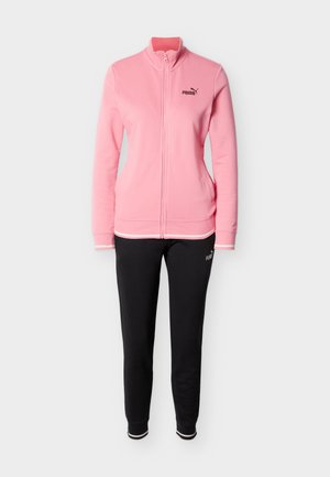 Rozā džemperis ar rāvējslēdzēju un augstu apkakli, uz kura ir melns logotips. Kombinēts ar melnām biksēm, abas izgatavotas no mīksta auduma un ar brīvu pieguli.