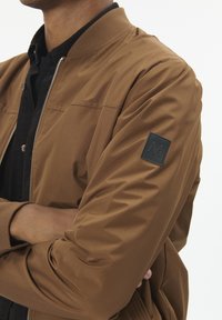 Chaqueta bomber marrón fabricada con tela lisa. Cuenta con un cuello acanalado y un parche de logo negro en la manga izquierda. Cierre frontal con cremallera.