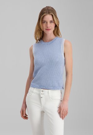 Frau trägt ein ärmelloses hellblaues Stricktop und weiße, hoch taillierte Hosen und steht vor einem schlichten grauen Hintergrund.