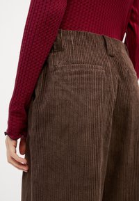 Brown corduroy trousers with vertical ribbing, featuring a back pocket and a high waist. Paired with a red and navy striped long-sleeve top.