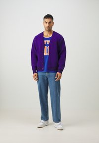 Lilla cardigan med sorte striber, knappet ned, båret over en blå basketballtrøje. blå jeans og hvide sneakers afslutter outfittet.