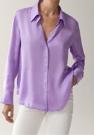 Frau trägt eine langärmelige, lavendelfarbene Bluse mit Knöpfen und Kragen, kombiniert mit weißen Hosen, Hand in der Tasche, vor schlichter Hintergrund.