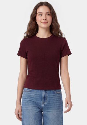 Junge Frau mit langen welligen Haaren, die ein schlichtes dunkelrotes Kurzarmshirt und blaue Jeans trägt, steht vor einem weißen Hintergrund.
