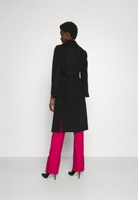 Schwarzer Trenchcoat mit einem taillierten Gürtel, langen Ärmeln und einem Schlitz hinten, kombiniert mit hellrosa Hosen und High Heels.