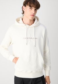 Sudadera con capucha color crema que presenta un bolsillo frontal tipo canguro, puños de canalé y el logo bordado de "Calvin Klein Jeans" en un beige más claro.
