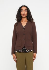 Brauner taillierter Blazer mit mit einem Reverskragen, zwei Fronttaschen und zwei Knöpfen. Über einer gemusterten Bluse und olivgrünen Hosen getragen.