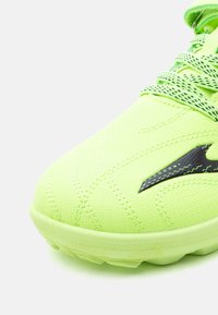 Κοντινή λήψη της μύτης ενός αθλητικού παπουτσιού σε νέον πράσινο, με λεπτομερείς ραφές, υφασμένο πέλμα, σχεδιασμένα κορδόνια και μαύρο λογότυπο της μάρκας.