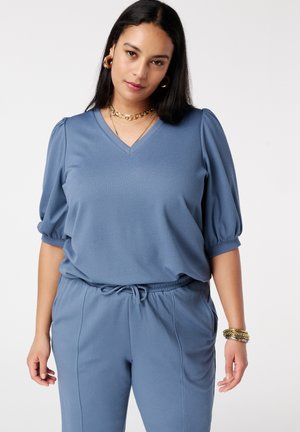 Femme portant un haut bleu texturé à col en V avec manches courtes bouffantes et un pantalon assorti à cordon de serrage, accessoirisée avec des bijoux en or.