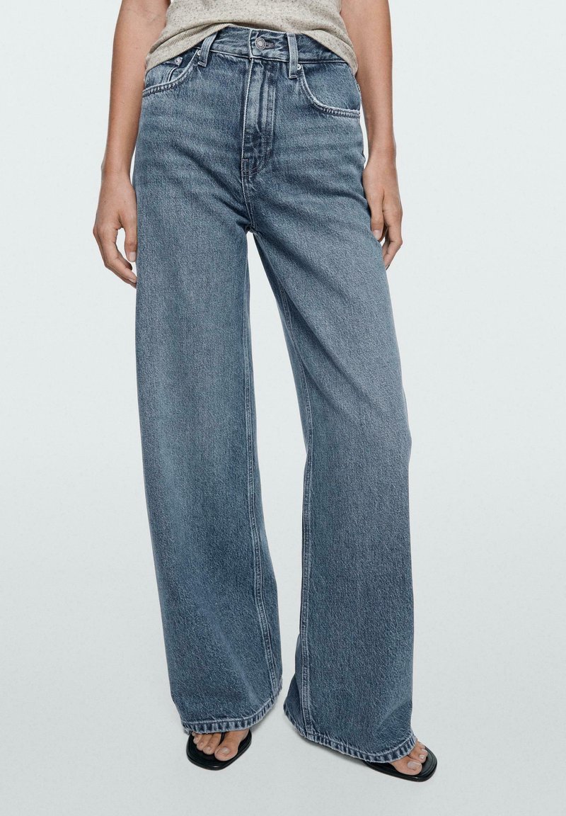 Persoon draagt een hoge taille, wijde blauwe jeans gecombineerd met een grijs topje en zwarte open tenen sandalen met een vierkante neus.