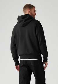 Sudadera con capucha negra hecha de tela suave, con un corte holgado y puños de canalé. Combinada con jeans oscuros, que muestran un estilo casual.