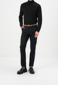 Schwarzer Rollkragenpullover, schwarze schmal geschnittene Hose, brafter Gürtel und schwarze Slipper mit strukturiertem Sohlen. Weißer Hintergrund.