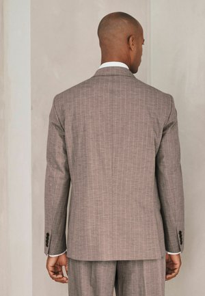 Borotvált fejű férfi világosbarna, tűselyem öltönyt visel, egy sima bézs fal felé áll, kezei lazán lógva oldalán.