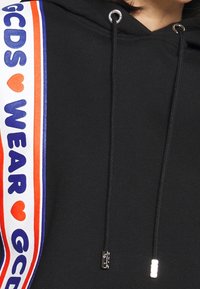 Sweat à capuche noir avec cordon de serrage, éléments en métal argenté et texte imprimé audacieux en bleu, rouge et blanc sur le côté. Texture de tissu lisse.