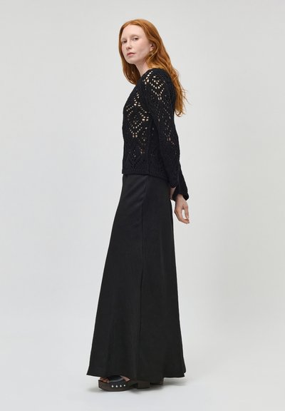 Femme aux cheveux roux portant un haut à manches longues en crochet noir, une longue jupe noire et des sandales plateformes noires, debout de profil contre un fond uni.