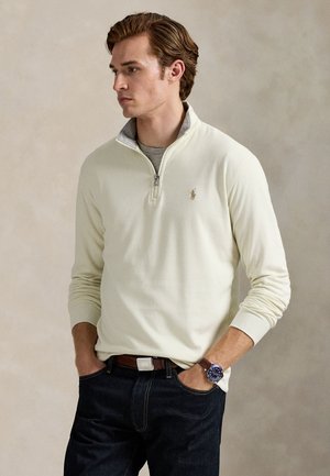 Jeune homme aux cheveux bruns portant un pull crème à fermeture éclair quart de zip, un sous-vêtement gris, un jean foncé, une ceinture marron et une montre bleue au poignet, les mains dans les poches.