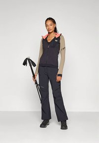 Egy nő kültéri dzsekiben és nadrágban, fekete túrbotokat tartva, egy sima világosszürke háttér előtt áll.