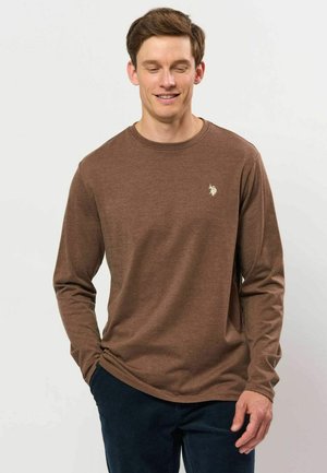 Ung mand smilende, iført en brun langærmet skjorte med et lille hvidt logo på brystet og mørke bukser, hænderne i lommerne.